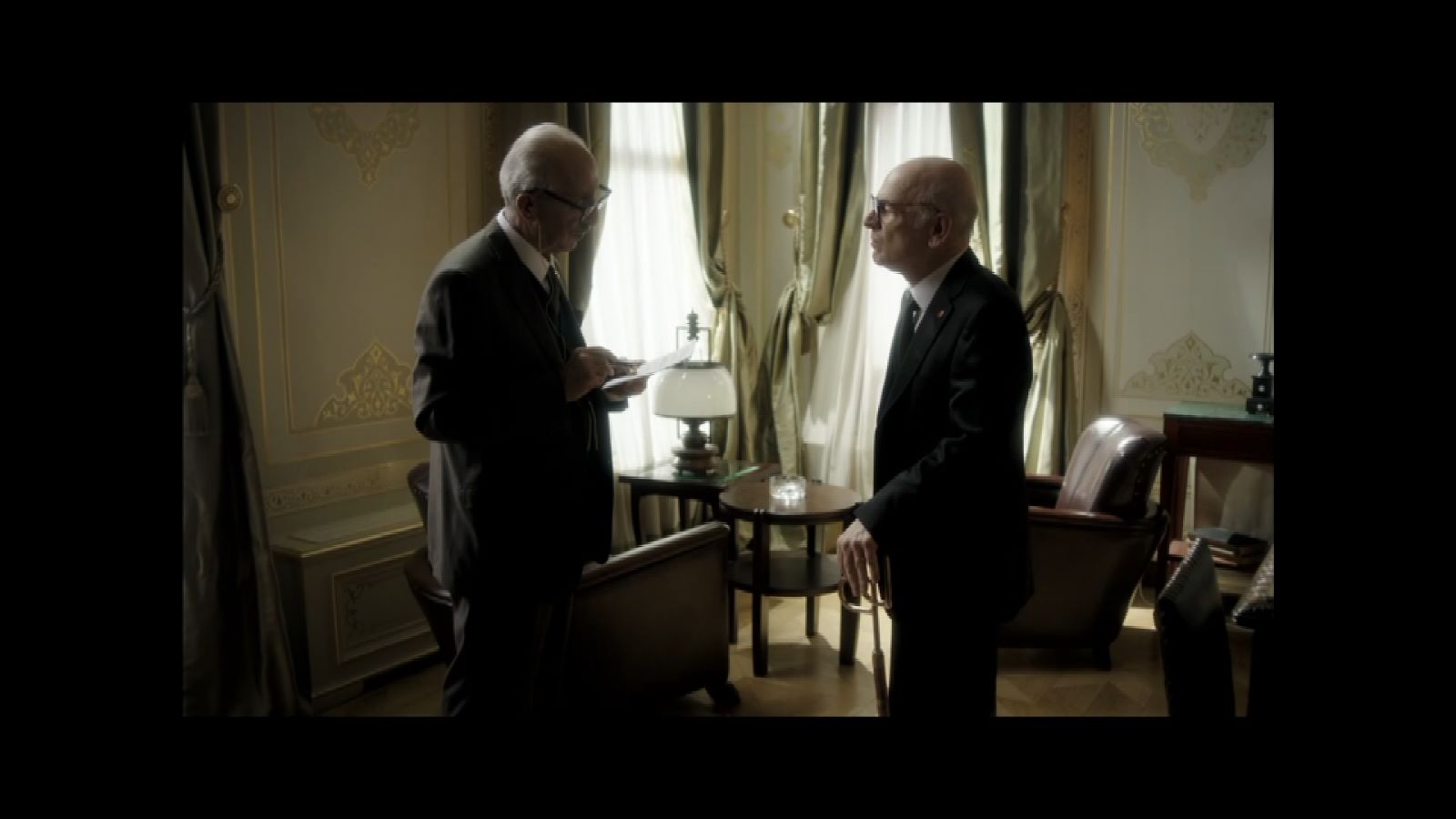 Ben Onu Çok Sevdim — Two suited men talking beside a bright window in a formal room.