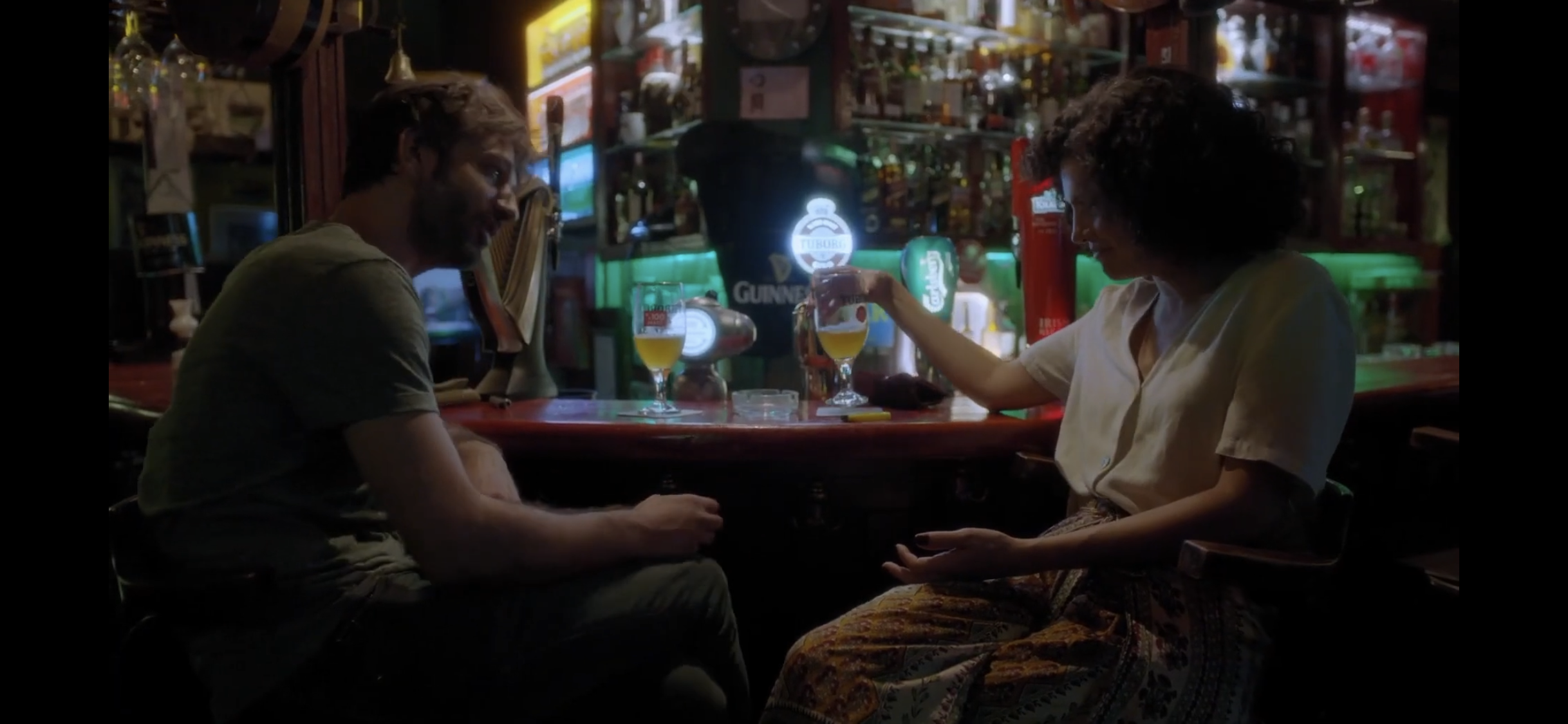 Dialogue — A man and a woman talking across a bar counter beneath colourful lights.