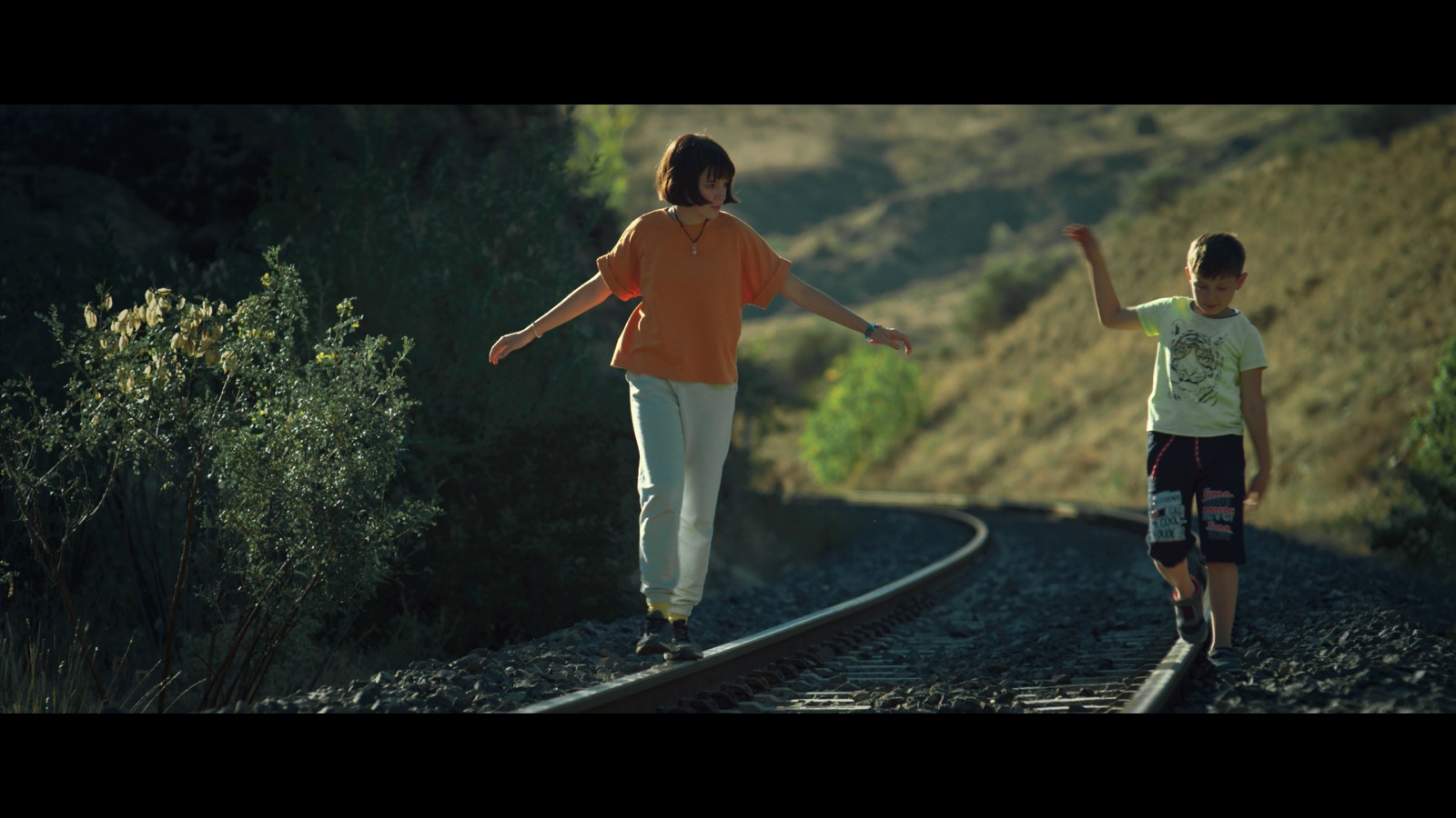Guilt — Two children balancing on railway tracks in afternoon light.