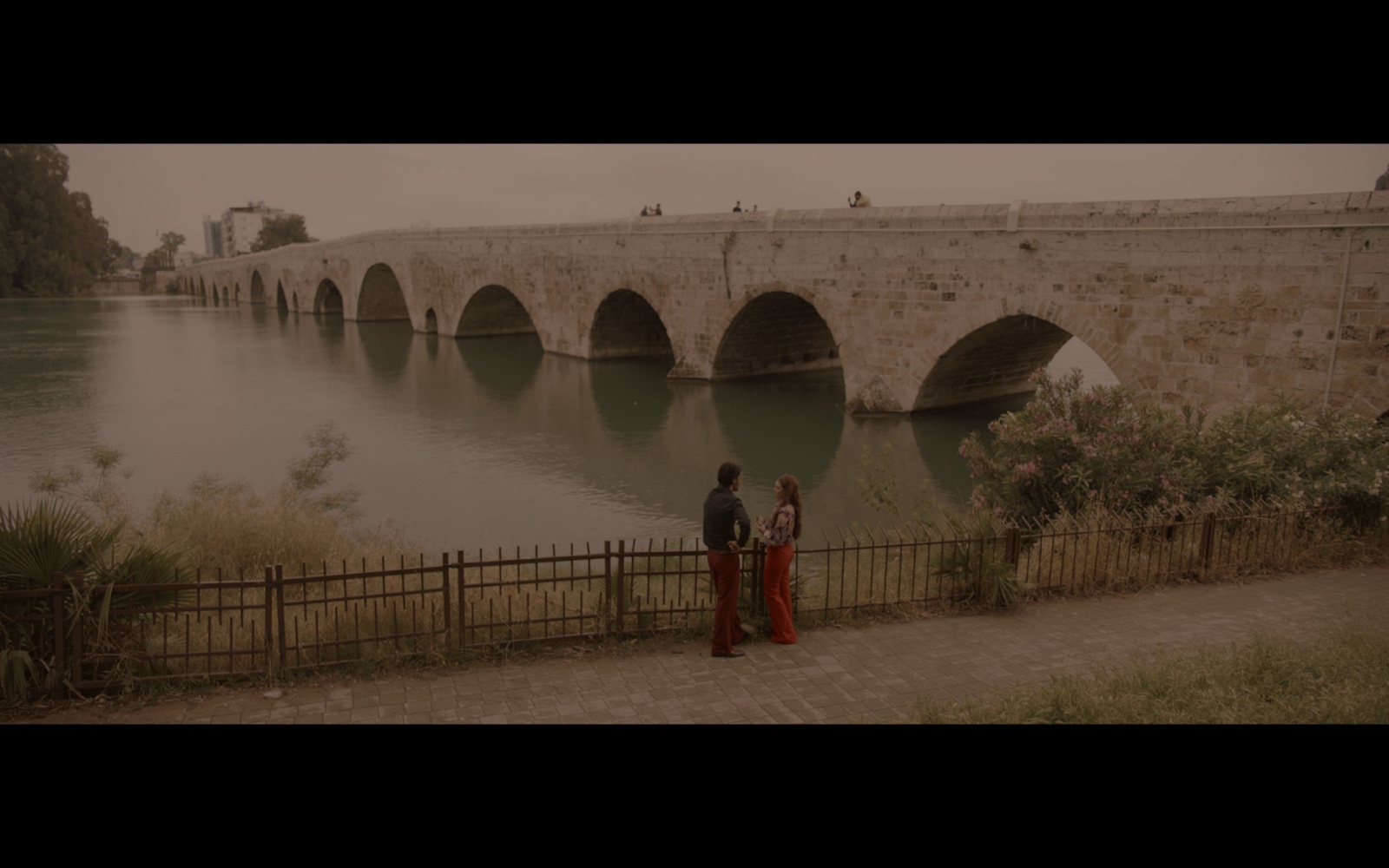 Radyo Günleri — Two figures beside a river and a long stone arch bridge.