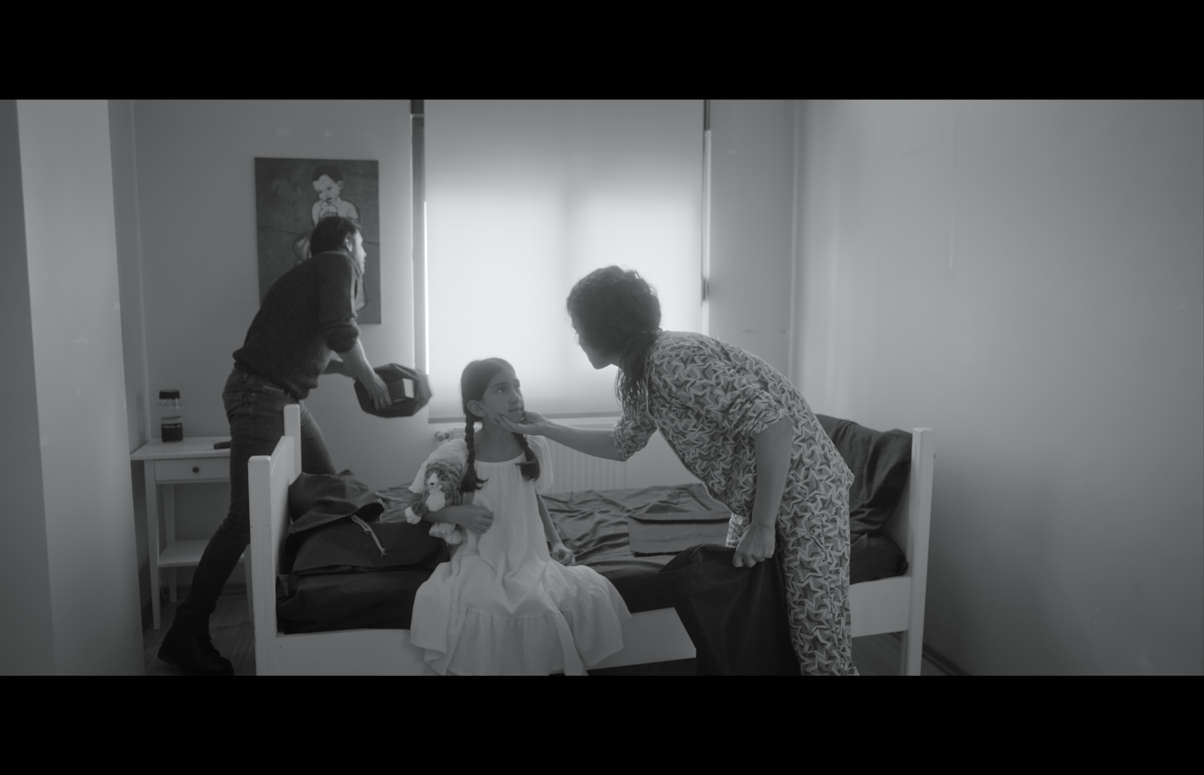 Sığınak — Black-and-white frame. A man and a woman leaning toward a girl seated on a bed in a bright room.