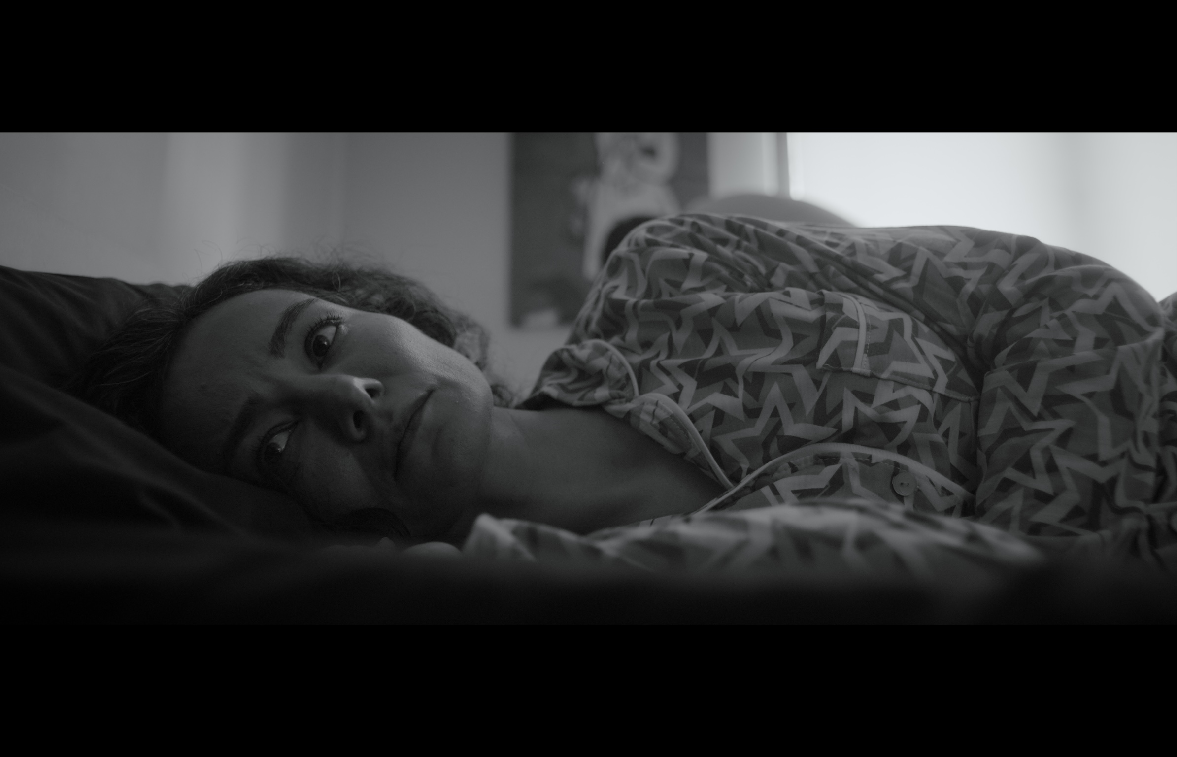 Sığınak — Black-and-white frame. A woman lying on a pillow in soft window light.