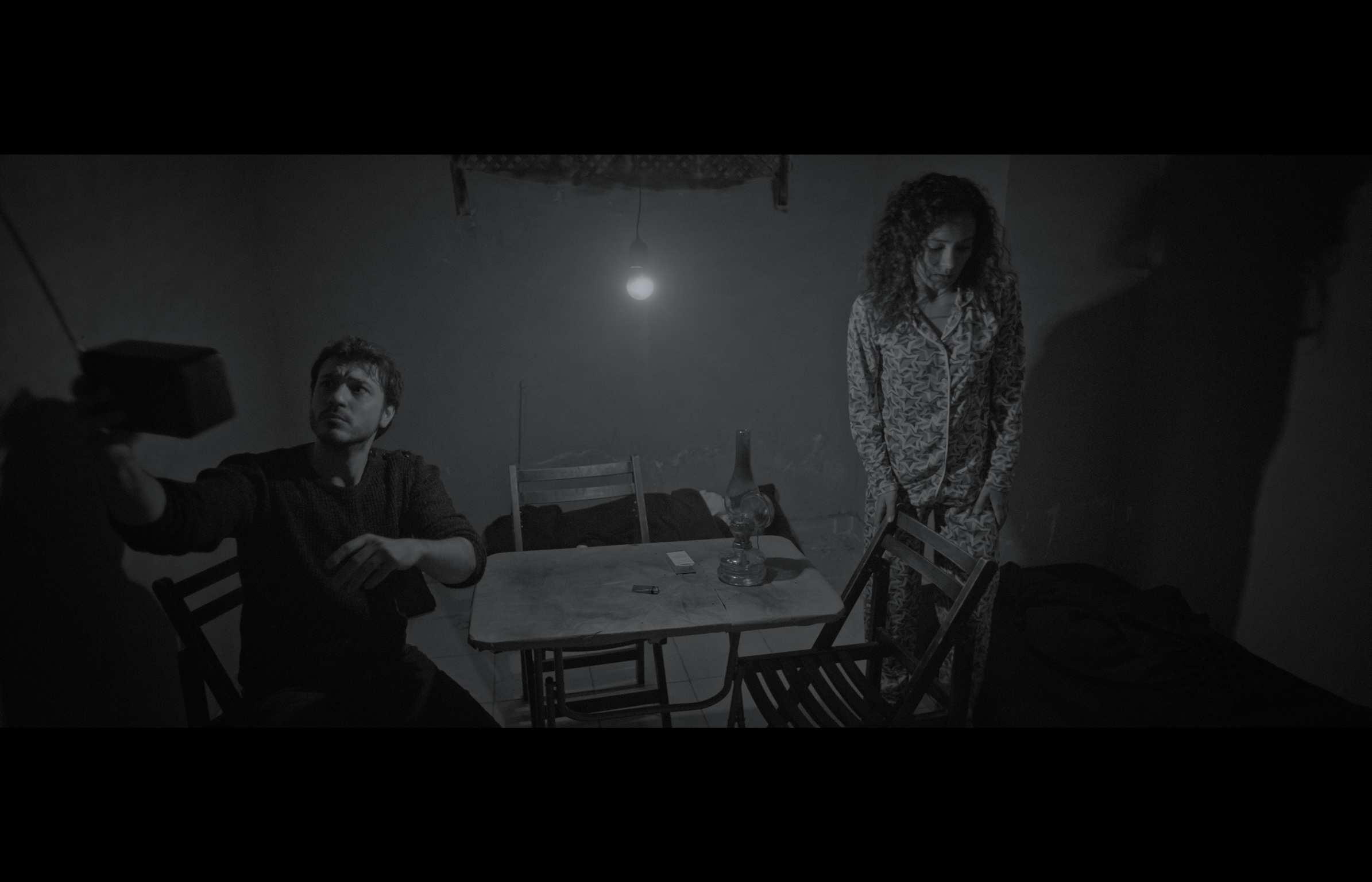 Sığınak — Black-and-white frame. A man seated beside a table and a woman standing in a dim room.