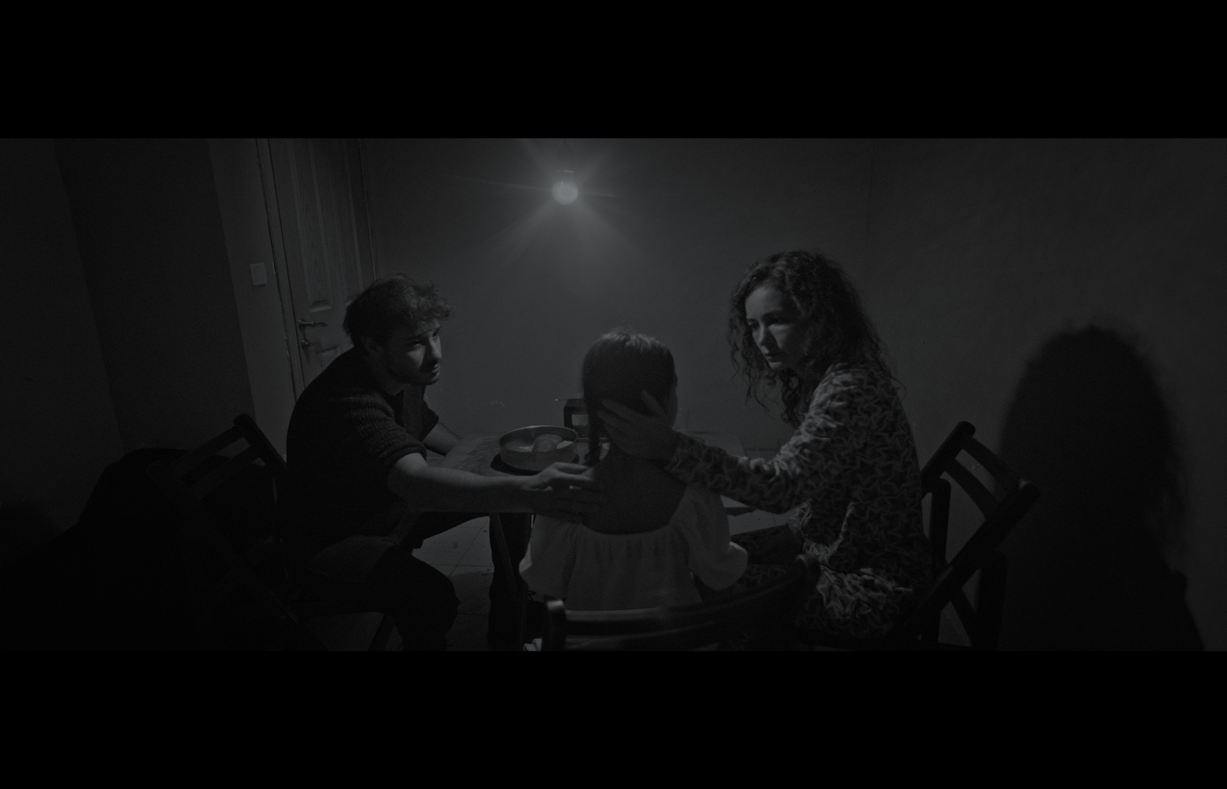 Sığınak — Black-and-white frame. Three figures gathered around a table beneath a single light.