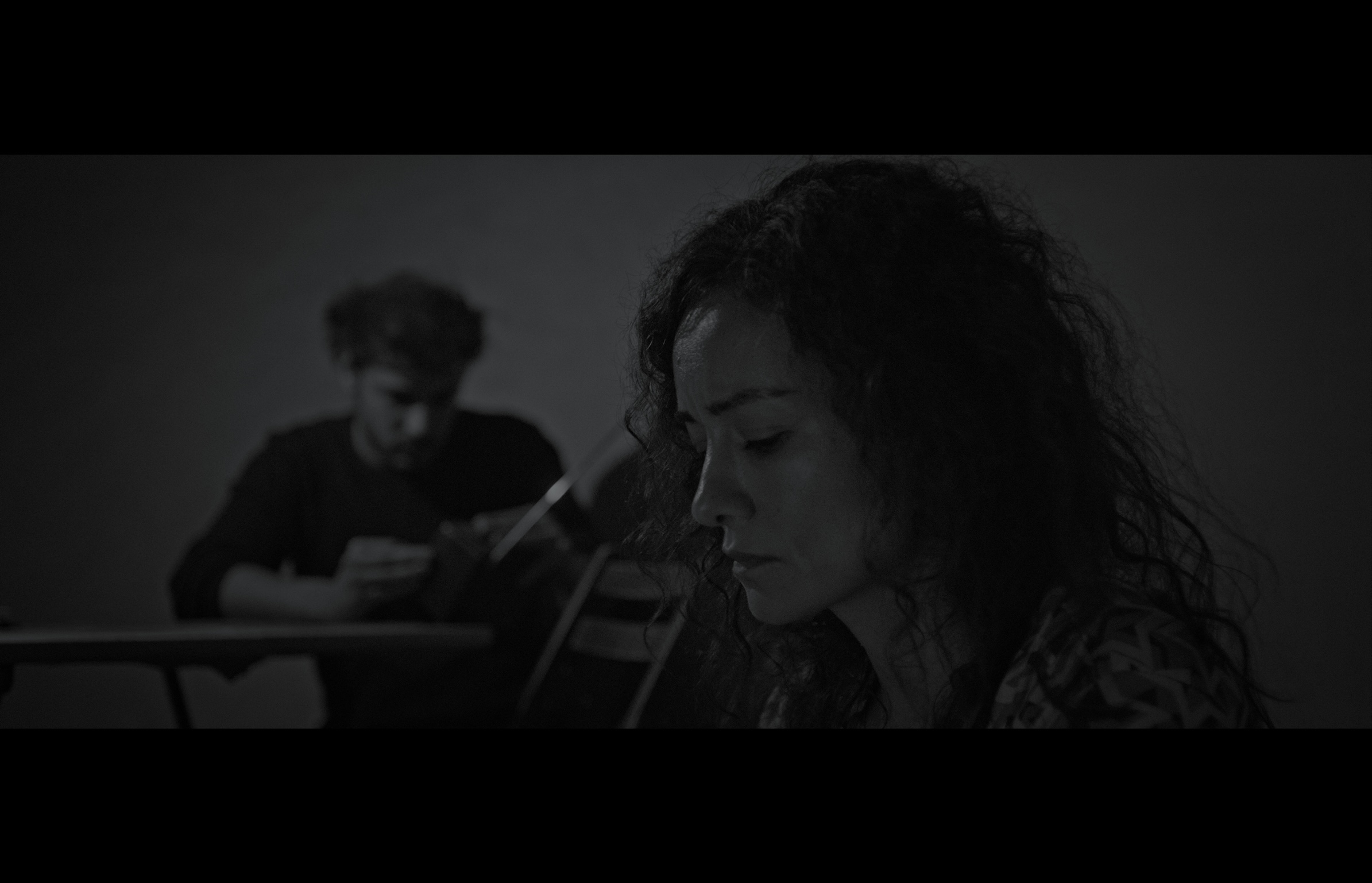 Sığınak — Black-and-white frame. A woman in the foreground with a man sitting at a table behind her.