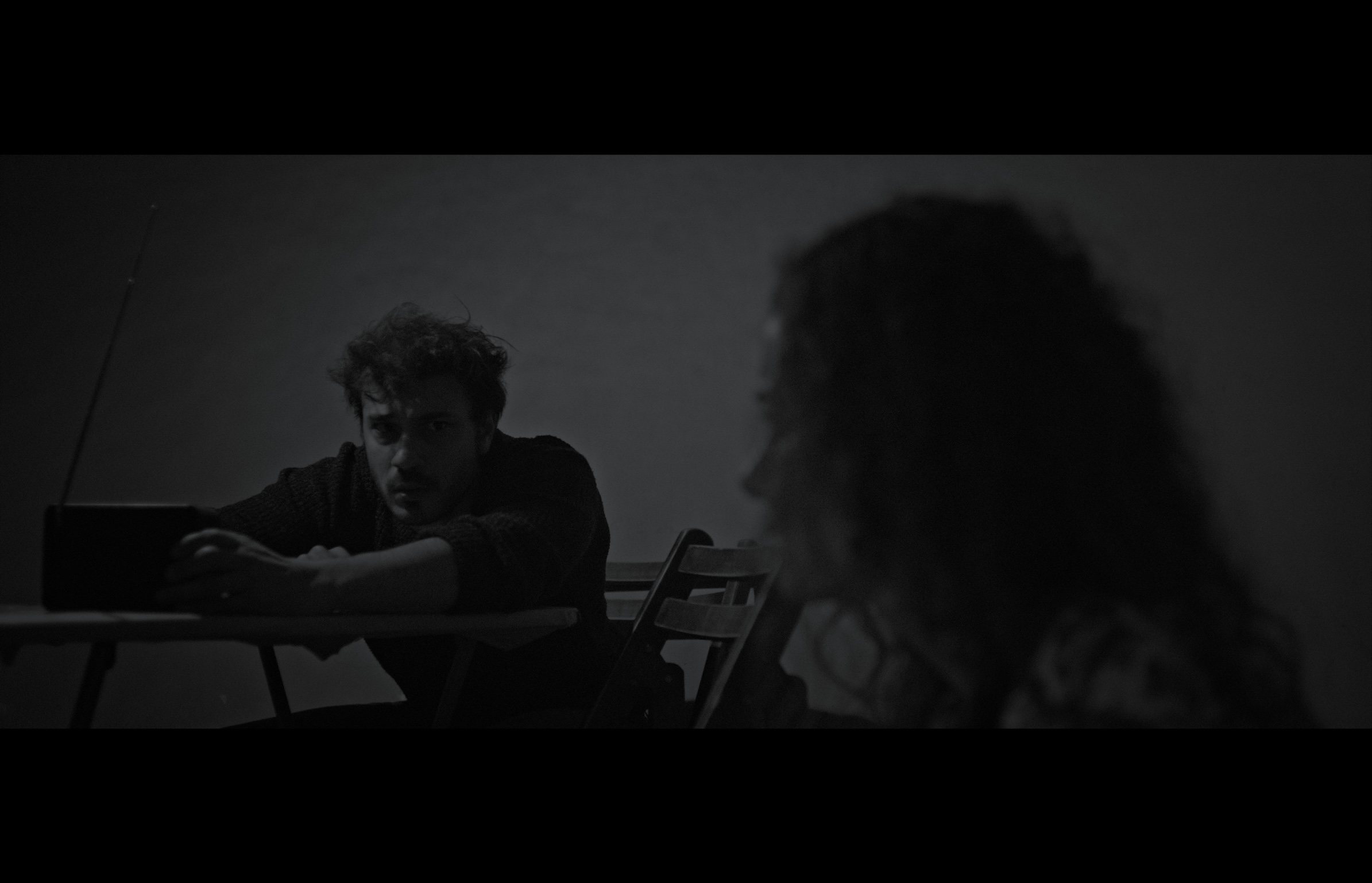 Sığınak — Black-and-white frame. A man leaning over a table, facing a woman in the foreground.