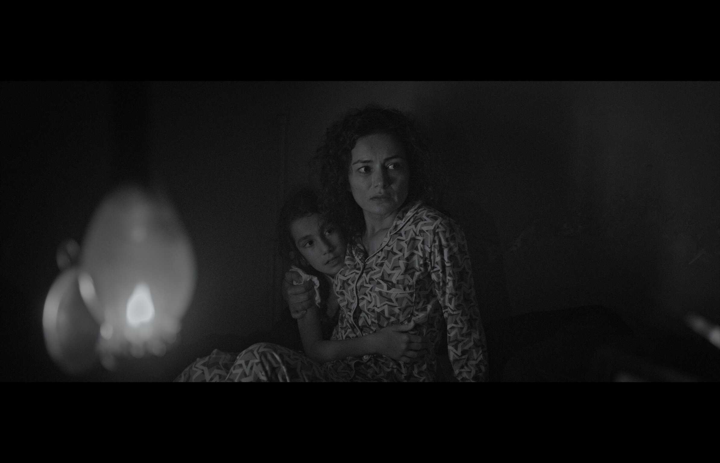 Shelter — Black-and-white frame. A woman seated beside the glow of an oil lamp.